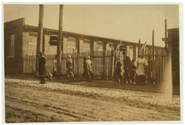 A sepia tone photo of ladies and men walking through a break in the fence.