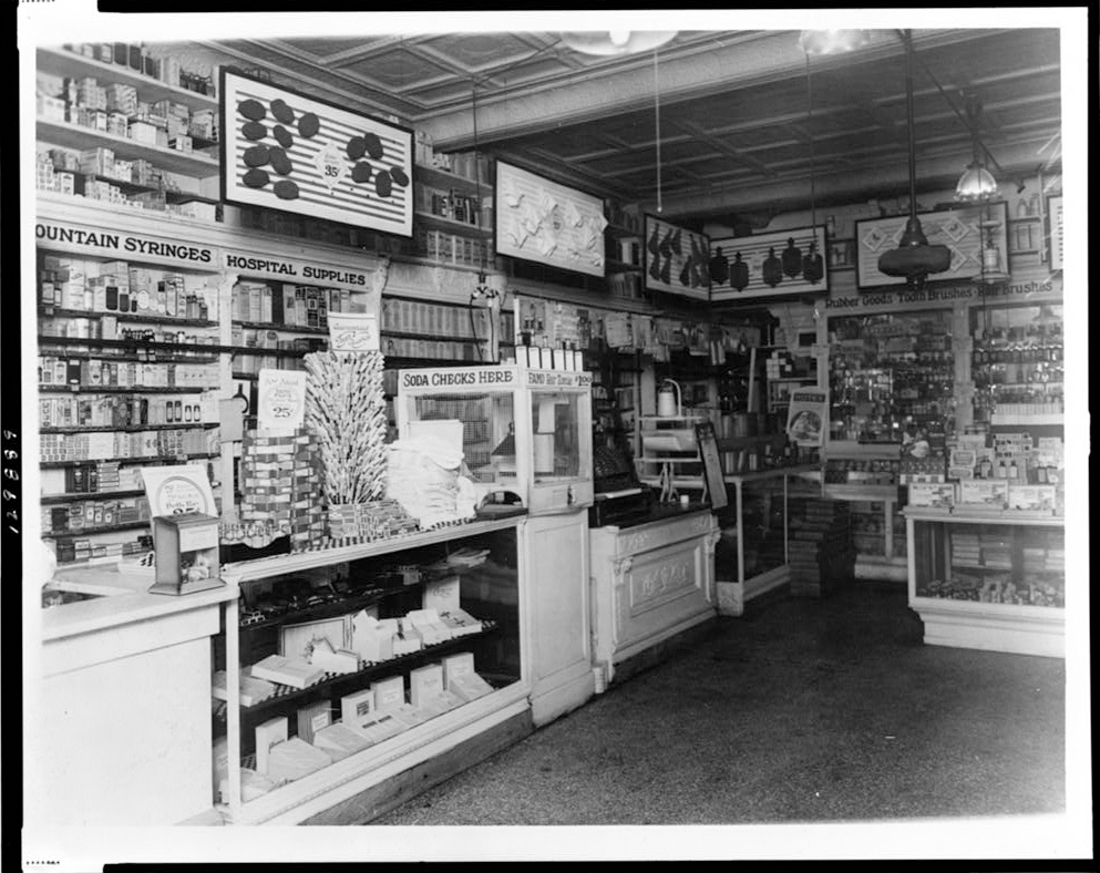 A black and white photograph of the interior of a drugstore.  Counters are stacked with goods and labeled “Fountain Syringes” and “Hospital Supplies.”