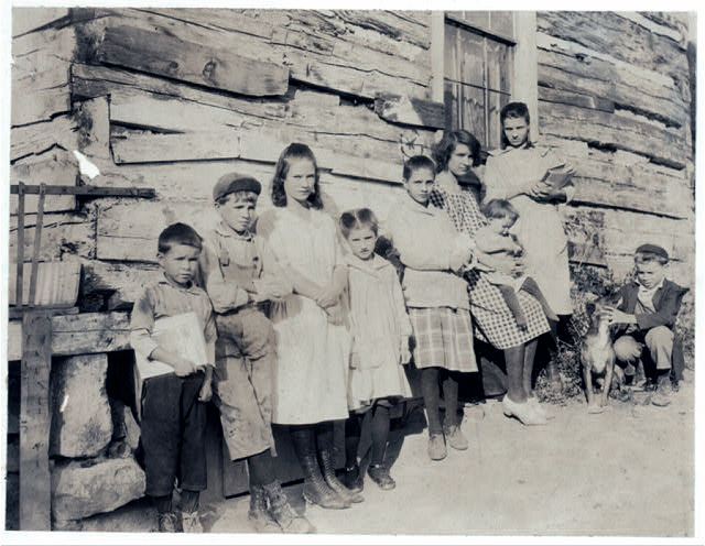 Black and white image of nine children ranging in age from about 1 to 16 years leaning against the side of a building, some carrying school books.