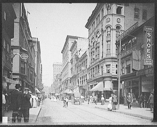 Numerous men and women go about their business in a busy street in c1906 Providence Rhode Island.  Several buildings and a horse-drawn carriage are visible in the background.