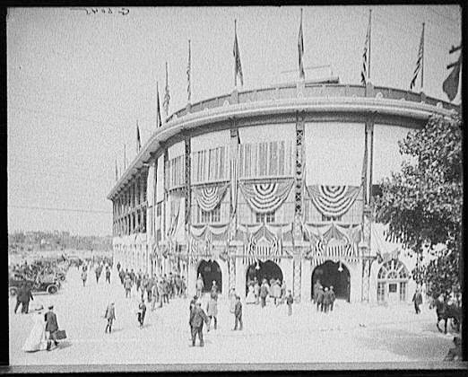 Dozens of men and women approach Forbes Stadium.  A few cars are parked to the left side.  Black and white, c1910.