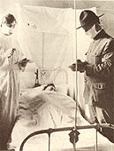 A military doctor and nurse wearing masks treat a sick patient who is lying in bed.