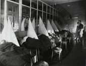 A hospital ward with beds which are draped in tents and sheets.  A nurse and a soldier are in the background.