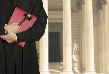 A judge holds his gavel and law book in front of a courthouse.