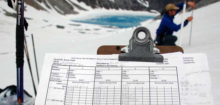 Photo of a glacier monitoring data sheet on a clipboard with a researcher in the background (Photo by Jim McLeod)