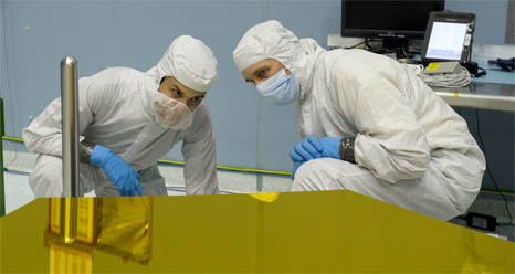 Two men in clean room suits look at a space telescope mirror