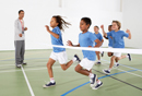 Photo of 5 children running through finished line ribbon, with adult coach timing them using a stopwatch