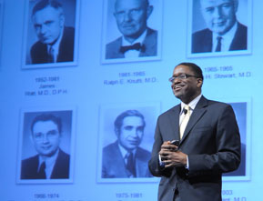 Photograph of Dr. Gary Gibbons giving a speech to the NHLBI community, with a digital presentation displayed behind him (showing the previous NHLBI directors).