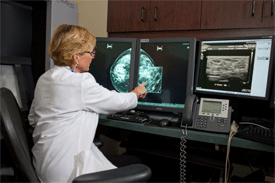 Blond woman physician in white lab coat examines two digital mammogram images on side-by-side flat screen monitors