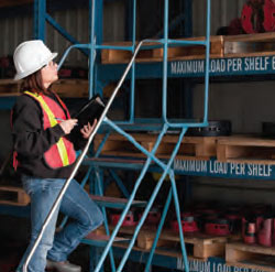 This is a photo of a woman wearing protective clothing; she is standing at the top of a ladder in a warehouse.