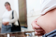 Photo of a woman looking at her reflection in a bathroom mirror and pinching her stomach fat.