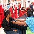 One young woman in a black v-neck shirt is having her blood pressure checked by a health care professional as she sits in front of a display of formal red dresses on mannequins.