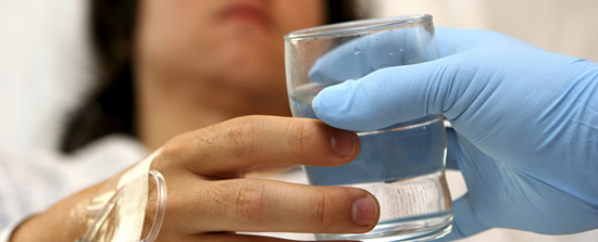 Female patient with a venous catheter taped down onto her hand; gloved hand or hospital worked handing her a glass of water.