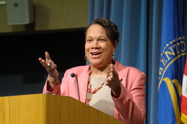 African-American woman in a pink blazer gives smiles and talks at a lectern