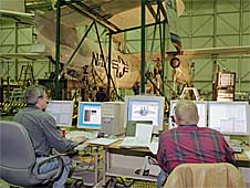 Two men sit at computers while testing an airplane in the background