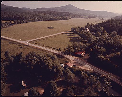 Rural scene showing the Naccochee Valley two miles southwest of Helen, Georgia, which is near Robertstown. The Chattahoochee River is visible at the bottom and top right in the photo. The red roofed building on the riverbank at bottom center is the old Nacoochee station, now a flower and crafts shop. The building across the road is an antique shop. The Nacoochee Indian mound is seen in the distant field beyond the junction of state highways 17 and 75, 05/1975 Documerica by Al Stephenson.