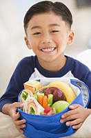 Photo: Boy with lunch box open. Contents are sandwich, apple, banana, carrot sticks and juice box.