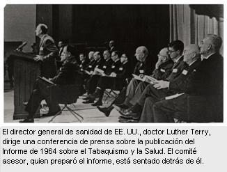 En esta foto en blanco y negro, el director general de sanidad, doctor Luther Terry, se dirige al público de la conferencia de prensa desde el estrado, (el público no aparece en la foto). Dos hileras de hombres están sentados en sillas plegadizas detrás de él. 