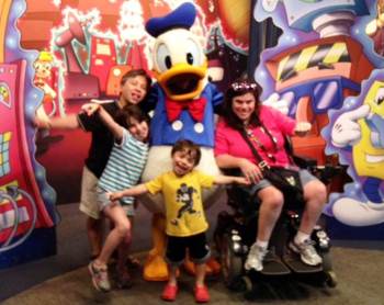 Image shows the Thomas family, a family of four. Posing for the camera with Donald Duck at Disney World.  All are facing forward. Looking jovial. Dan, the father, is on the left side of Mr. Duck with his right hand sticking out to the side. He is sticking out his tongue partially in a joking manner.  Abigail, the seven-year-old daughter, is standing in front of Dan, leaning on Mr. Duck, with her right arm out to the side and her tongue sticking out as well.  Noah, the seven-year-old son (Abigail&amp;#8217;s twin brother), is standing in the center of the image directly in front of Mr. Duck, smiling, with both arms outstretched beside him.  Jennifer Thomas, the mother, is sitting in her power wheelchair, to the right of Mr. Duck, leaning on him with her right hand in Noah&amp;#8217;s and her left hand to the side facing forward.  The background is a colorful Disney scene.