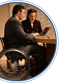 Photo of a man in a wheelchair sitting at a table working on a laptop computer. A woman is sitting next to him.