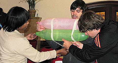 A man holds two long balloons while two other people tape the balloons together