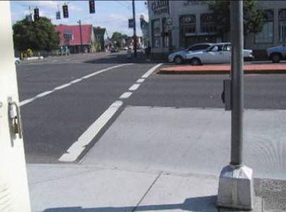 Photo of crossing, looking across wide street with a raised median island, with parallel street on photographer’s left.  Pushbutton is on a pole that is 8 to 10 feet outside the crosswalk lines.