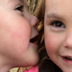 photo of a 3 year old girl whispering into the ear of her twin sister.