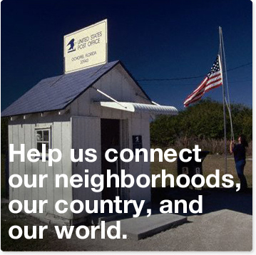 Help us connect our neighborhood, our country and our world. A small United States Post Office, about the size of a shed, with a blue roof and white walls in a secluded rural area. A woman is standing just outside the office, raising a United States flag on a mast.