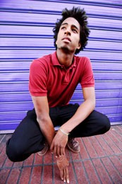 A young African American man crouches on a sidewalk and looks up.