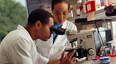 An African American male researcher looks through a microscope as an African-American female researcher looks on.