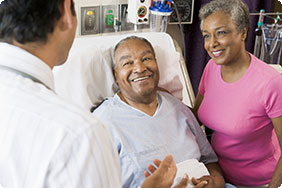 Older Man in a Hospital Bed and Older Woman Standing Beside Him Smiling at Doctor