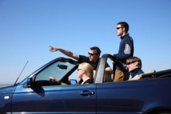 Photo shows an adolescent girl driving a convertible with three other adolescents as passengers—one has his right arm extended above the windshield, pointing at something in the distance, and another is standing in the back.