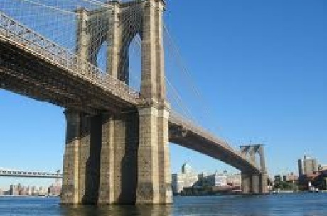 Under side view of a bridge extending out over water. Blue skies above and city in the background. 