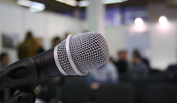 Focus on a microphone with a blurred mingling audience in the back, signifying "public comment"