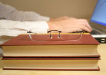 A pair of glasses rest on a research book in front of man on a computer