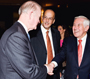 a photo of three men standing together; two shaking hands.