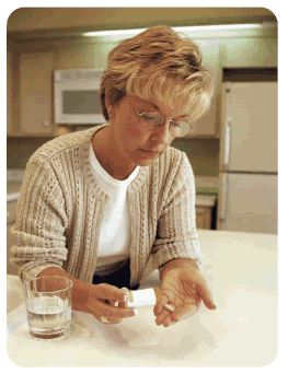 Photo of blonde woman seated in her kitchen with a glass of water and a pill bottle.