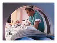 Photo of a patient lying under the circular opening of an MRI machine. The patient’s legs and feet are inside the machine. A health care provider attends to the patient, standing by her upper body outside of the MRI machine.  