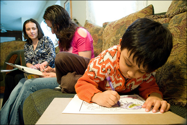 A 3-year-old boy colors in a book on a couch while a Promoting Positive Parenting Program coach helps his  mother fill out a survey.