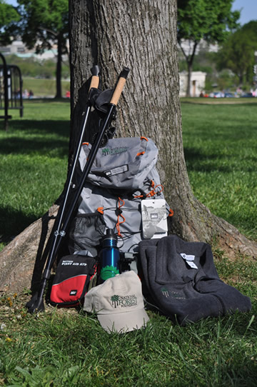 a daypack, a set of trekking poles, a fleece vest, a National Forest Foundation baseball cap, a water bottle, a first aid kit all set up against an urban tree.