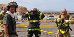 A structural firefighter in a helmet looks at the camera, while other firefighters nearby don structural firefighting gear.