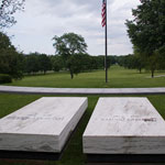 Two marble grave stones and a flag overlook a park vista.