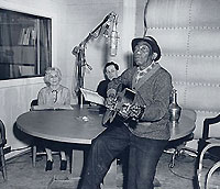 Mississippi John Hurt playing a guitar,  with Rae Korson and Joseph C. Hickerson in the background