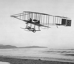 Black and white photo of a flying machine.  The pilot is completely exposed to the elements, and the plane is a frame with no walls.