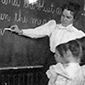 Elementary School Children Standing and Watching Teacher Write at Blackboard