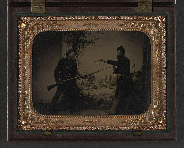 [Two unidentified soldiers in Union uniforms posing with bayoneted Springfield Model 1861 rifled musket with attached bayonet,  knife, and Colt Model 1851 Navy revolver in front of painted backdrop showing landscape with camp] (LOC)