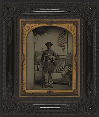 [Unidentified African American Union soldier with a rifle and revolver in front of painted backdrop showing weapons and American flag at Benton Barracks, Saint Louis, Missouri] (LOC)