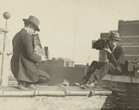 Two photographers taking each others' picture with hand-held cameras while perched on a roof