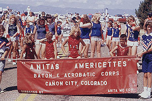 Young girls in costume wait their turn to participate in the parade, holding a banner that reads, "Anita's Americettes Baton and Acrobatic Corps, Canon City, Colorado." 