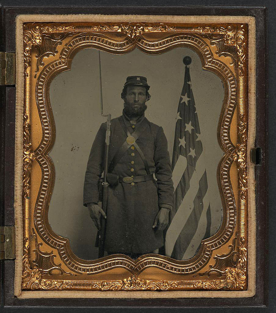 [Unidentified soldier in Union uniform and Company H cap with bayoneted musket, cap box, and Volunteer Maine Militia (VMM) belt buckle in front of American flag] (LOC)
